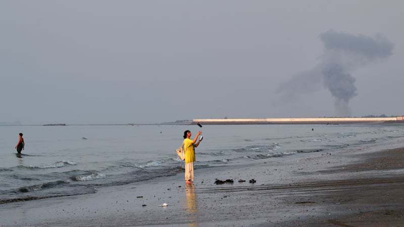A woman stands at the water's edge along the Strait of Hormuz as a plume of smoke rises in the...